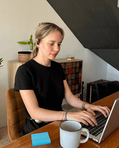 alt="Femme portant les écouteurs traducteurs Hearific en visioconférence depuis son ordinateur portable, dans un cadre de travail à domicile"